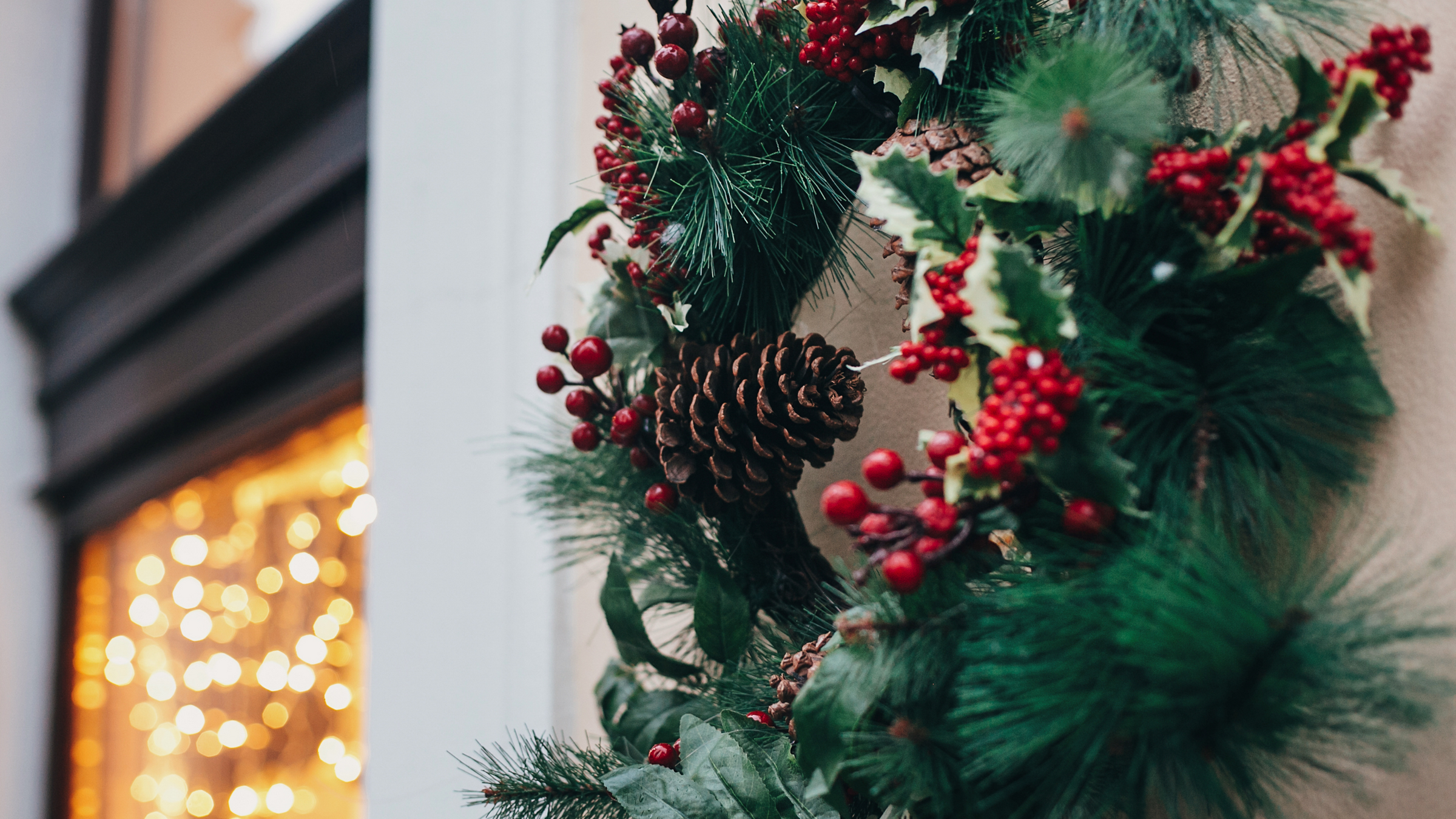 pine cone and berry wreath hanging on a wall with warm white lights in the background