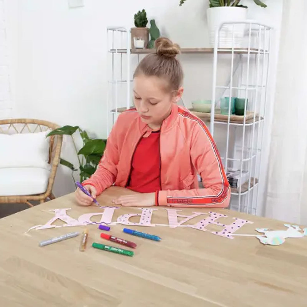 Young person at a table using glitter glue pens to create a happy birthday banner