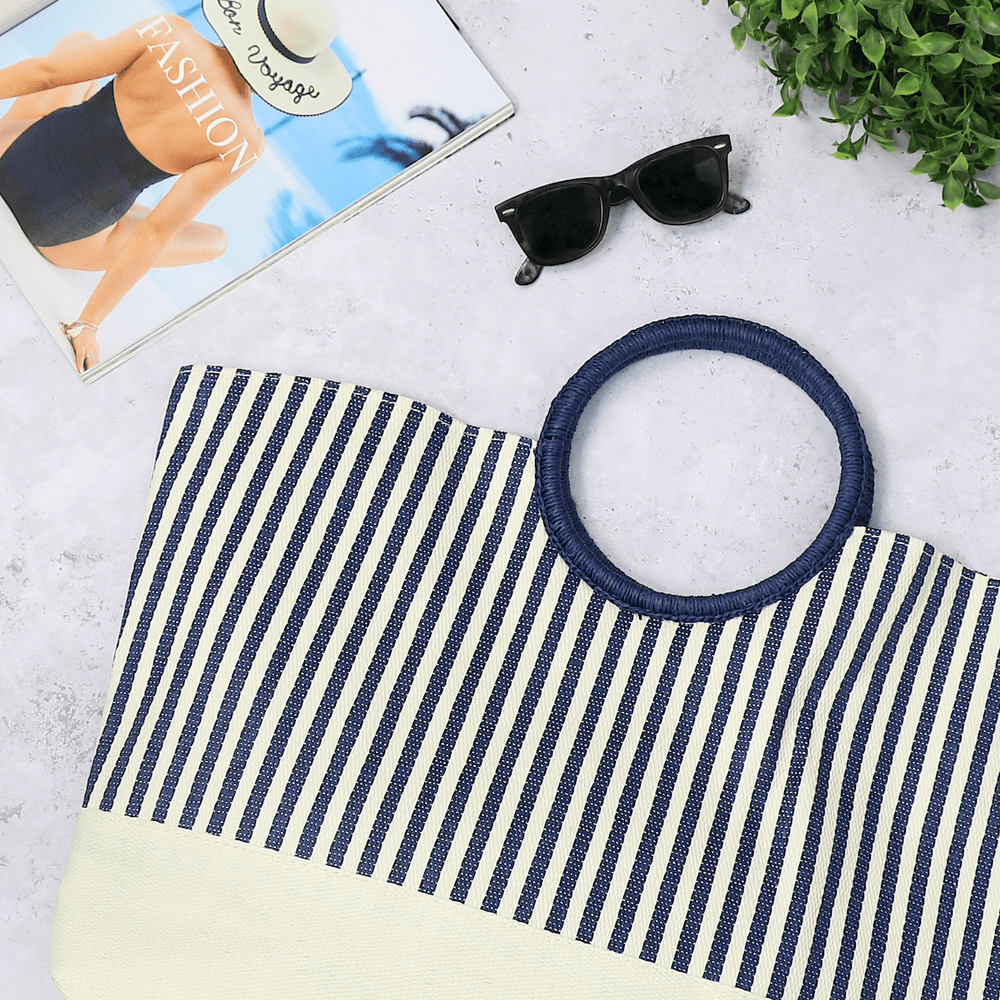 blue and cream striped beach bag on concrete background beside magazine, sunglasses and green foliage