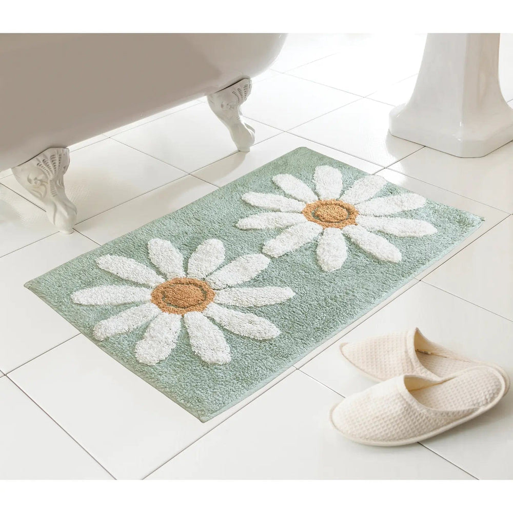 Teal green colour bath mat with 2 large white daisies pattern on a tiled floor next to a bath and slippers