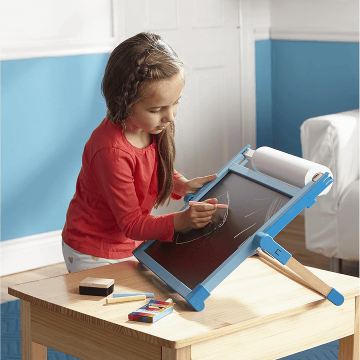 little girl uses red chalkstick to draw a picture on the chalkboard resting on wooden table