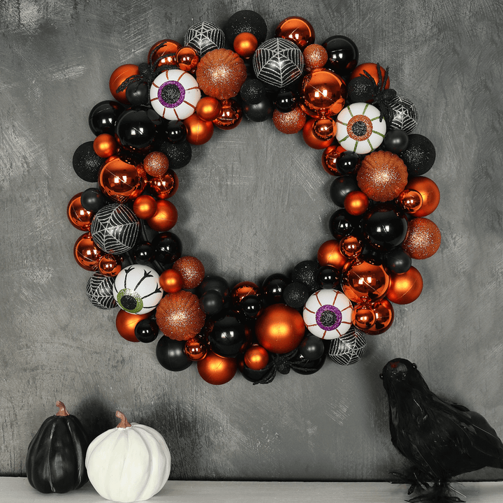 halloween wreath featuring orange and black baubles with eyeball glitter baubles hanging on black wall above a fireplace ledge featuring black crow decoration and white and black pumpkins