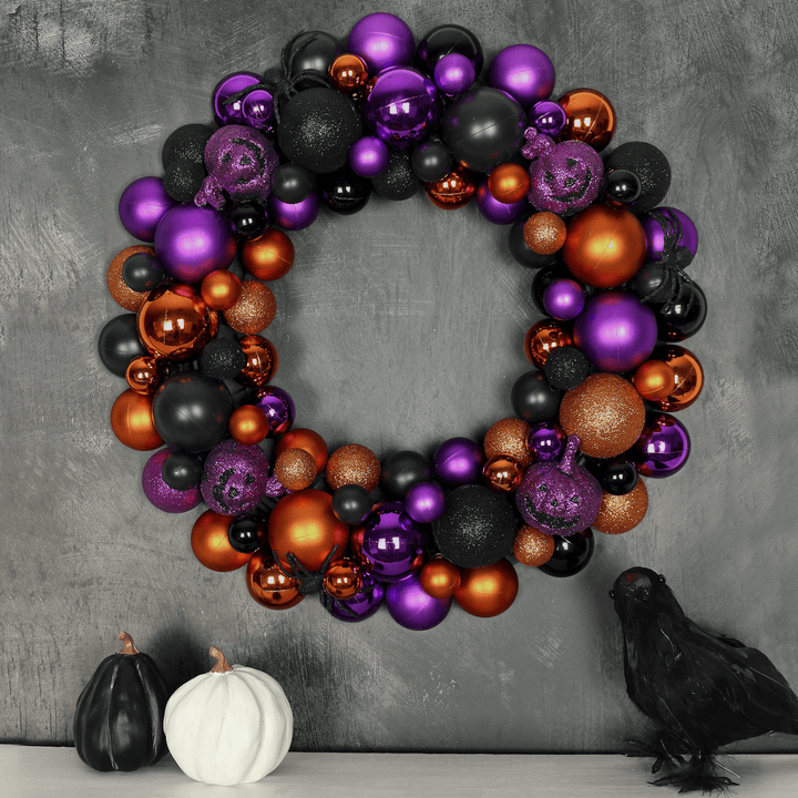 Purple black and orange halloween bauble wreath on a black background above a fireplace ledge featuring white and black pumpkins and a crow decoration