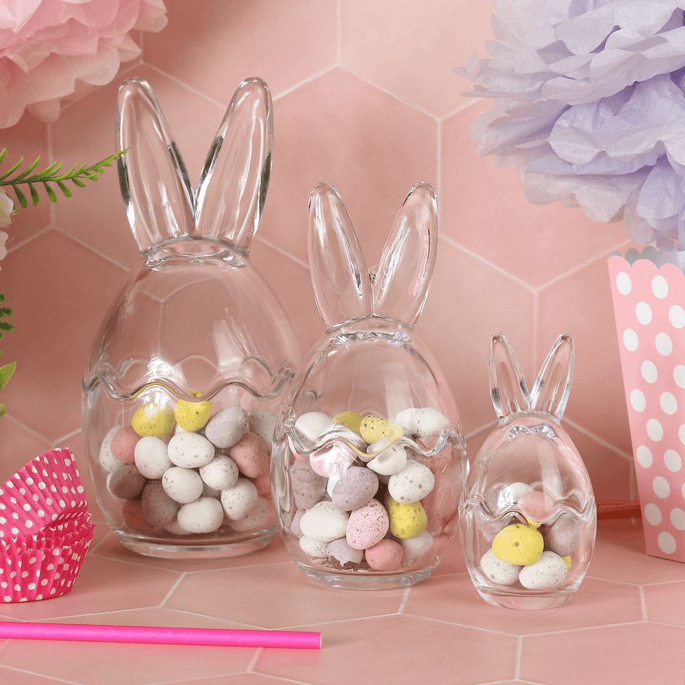 set of 3 bunny ear glass storage jars filled with mini eggs on pink background with cupcake cases and boxes to the side